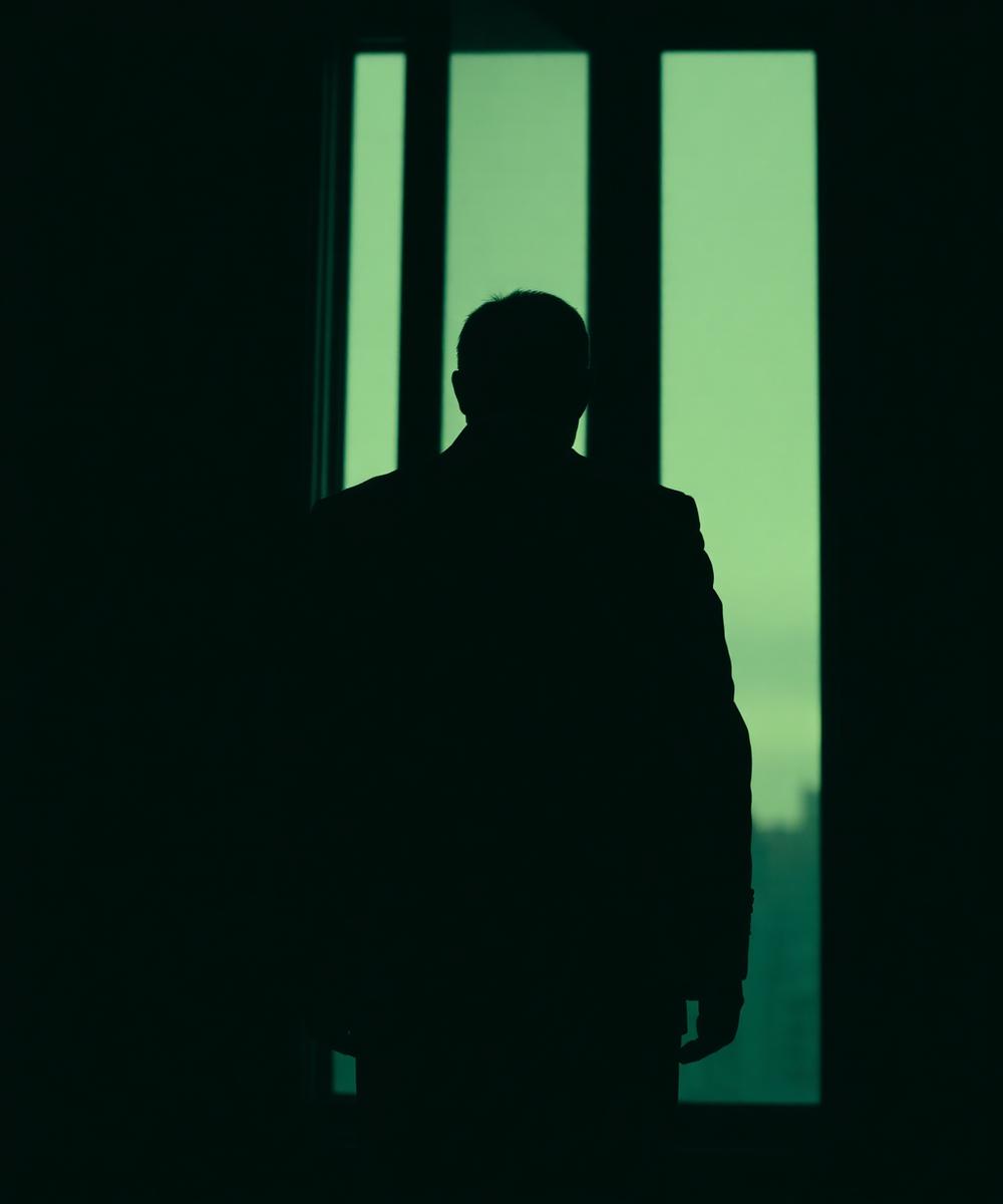 Silhouette of a man in a tailored suit standing before a tall office window at dusk